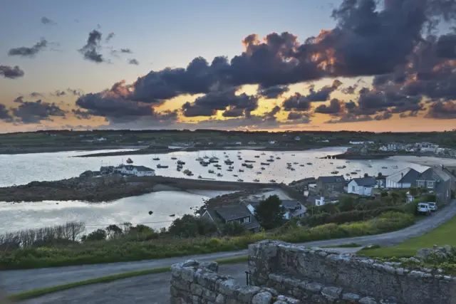 The view across Hugh Town on St Mary's on a gloomy day. There are boats anchored in the bay and some houses dotted along a road.