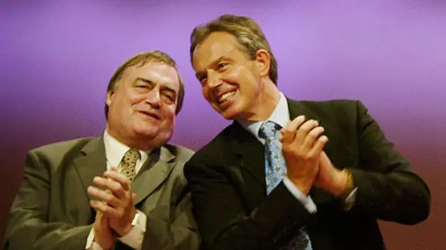 John Prescott (L) leans in to speak to Tony Blair (R) as they both clap at Labour conference in 1998. Prescott is in grey suit, white shirt and patterned tie while Blair is in black suit, light blue shirt and patterned tie