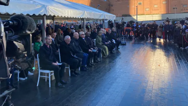 Guests sit under a marquee against the reflective background of Birmingham New Street station. The Odeon building can be seen in the background. Onlooks surround a cordon.