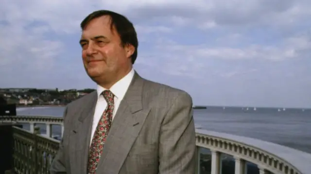 John Prescott posing for photo in light grey suit, white shirt and patterned tie in front of a beachfront view