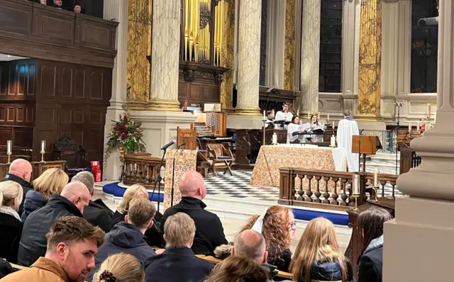 A congregation assembled in pews in a Cathedral. The altar can be seen, with priests and singers at the front. There are tall stone pillars and the altar is covered with a patterned cloth.