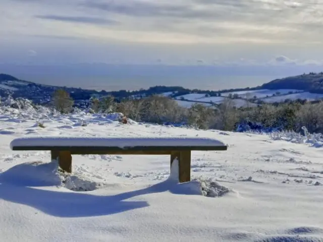 A wooden bench covered in snow, with heavy snow on the ground overlooking snow covered fields