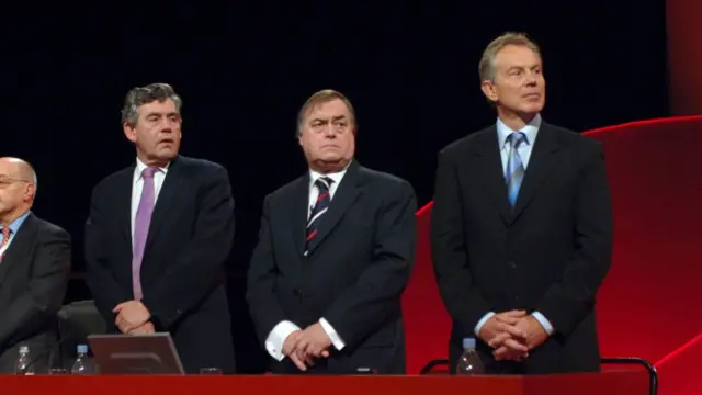 Brown, Prescott and Blair stand in a row behind a desk which says ‘Labour’ on it and has a rose. The three men wear suits and have their hands crossed in front of them.