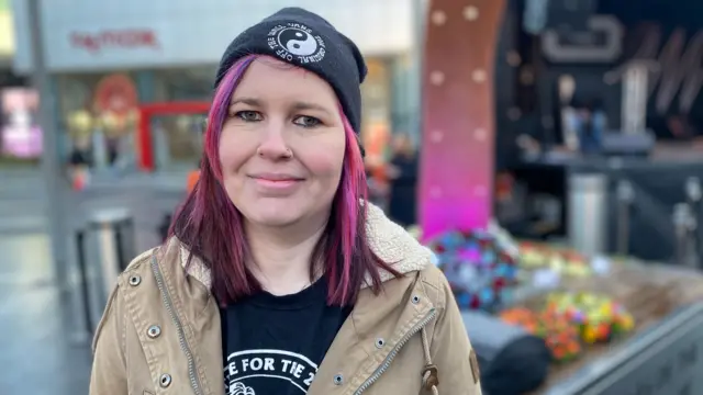 Amy Hodgins at the service in Birmingham. There are flowers in the background behind her. She has pink hair and is wearing a hat.