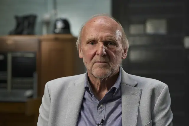 A man in a grey suit jacket and blue shirt. He is balding, with white hair. He sits in a room which appears to be a police station, a police helmet visible out of focus on a cabinet behind him.