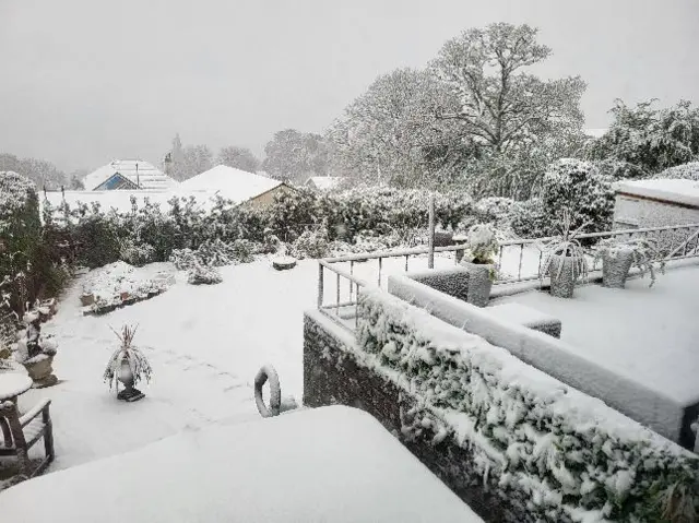 The photo shows a garden in Sidmouth that has been covered in snow. The garden's decking is to the right of the photo with some steps leading down to the grass.