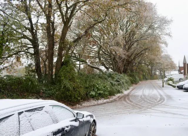 A snowy road in Woodbury. A car, covered in snow, is parked next to the road. Tyre marks cover the snow covered road.