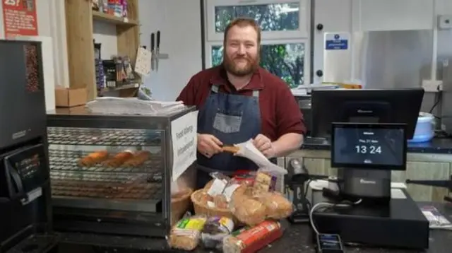 Shop Manager Dan stood behing the counter in an apron, putting a pasty in a bag.