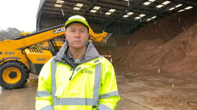 Mr Bayley stood in front of a large pile of salt. There is a digger behind him. He is wearing a high visability jacket and a hard hat