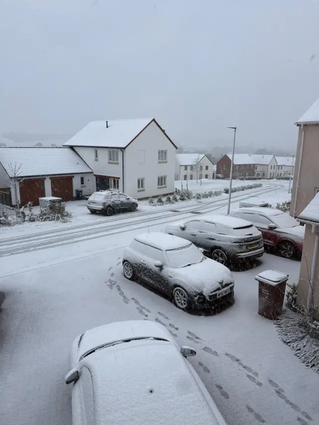 Snow has settled on a residential street. Cars are covered in snow. Footprints are in the snow.