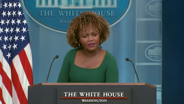 Karine Jean Pierre stands at the White House press briefing podium and wears a green dress