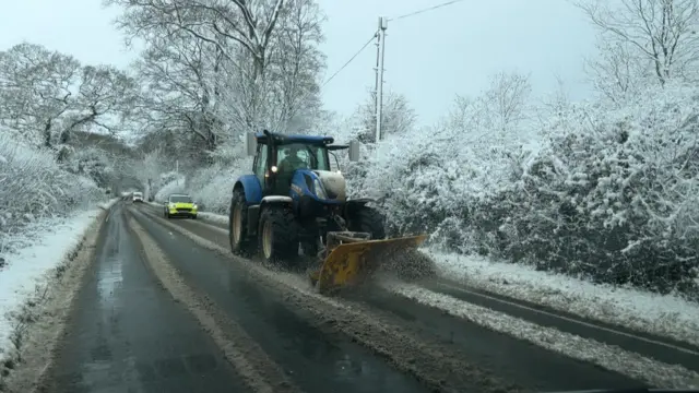 A tractor with a snow shovel on a road clearing snow with a police car behind.