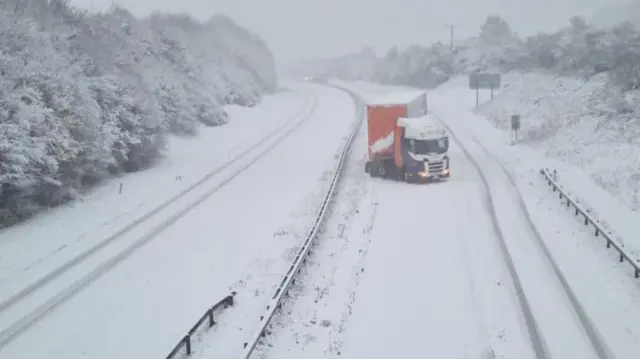 The image shows a stranded lorry on the A30 near Whiddon Down, Devon. The lorry has stopped with the cab facing the wrong way. Both carriageways have been covered in snow.