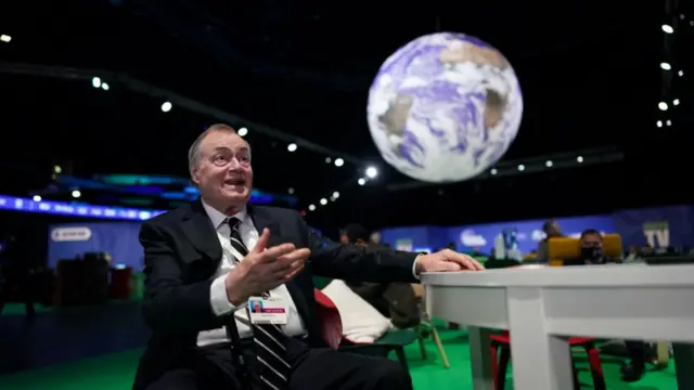 Long-shot of John Prescott at COP26. He's sitting down with his left hand resting on a white table to his side. He's wearing a black suit, white shirt and black striped tie, a cane resting on his right arm. In the background is a model of planet Earth