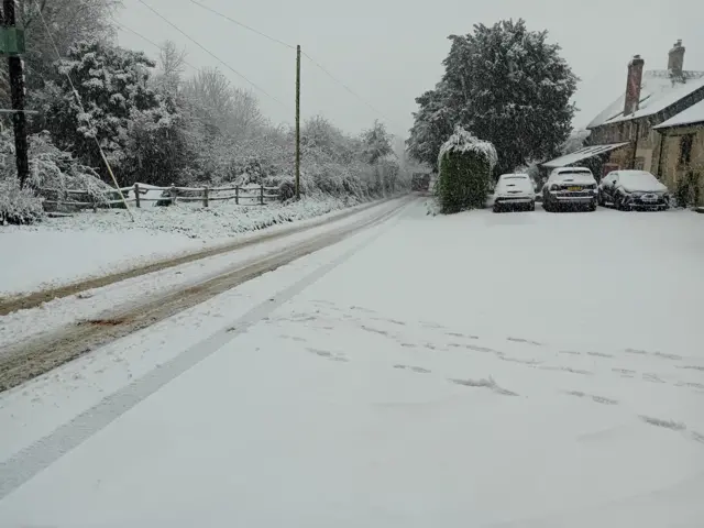 A road is covered in snow with tyre tracks. Cars are under a blanket of snow