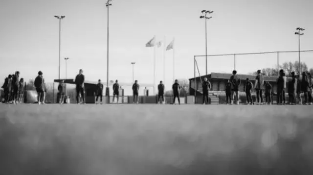 A black and white image of people standing still on a football pitch. There are about 23 people in the picture, dressed in tracksuits, standing with arms behind their backs. The photo is taken from ground level and floodlights, stand and tall flags can be seen.