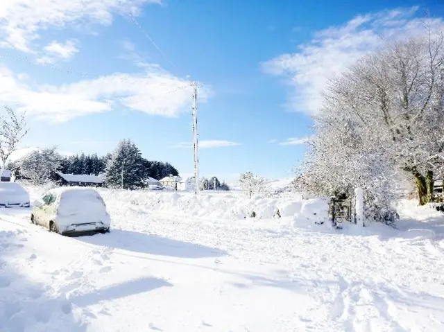 Heavy snow covers the ground, cars and trees with a blue sky