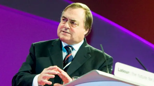 John Prescott speaking on stage behind a lectern. He's in dark suit, light blue shirt and striped tie looking to his right, his hands extended in front of him, fingertips touching