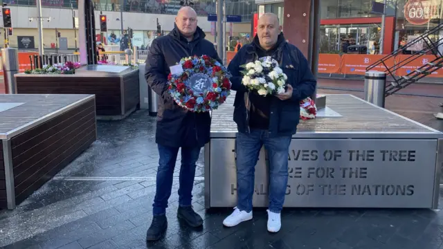 Matt Glover and Glenn Randall holding wreaths. The pub bombings sculpture is behind them.