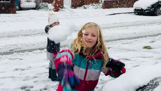 Two children playing in the snow in the street. One child in a brightly coloured winter jacket is throwing a snowball at the camera