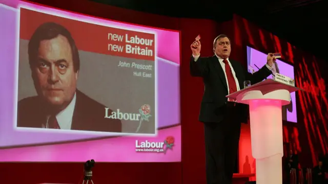 ohn Prescott holds up a couple of Government pledge cards as he delivers his closing speech at the Labour Party Annual Conference in 2004. He's standing behind a lectern in black suit and red tie. Behind him a screen is projecting a photo of his 1997 electoral pledge