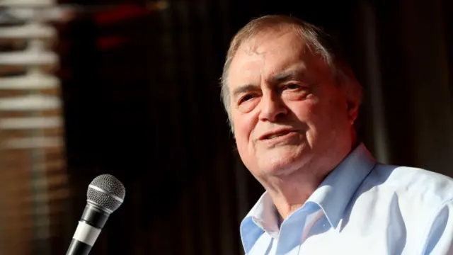 John Prescott speaking on stage. He's in a light blue shirt unbuttoned at the top, a mic in front of him as he looks to his left