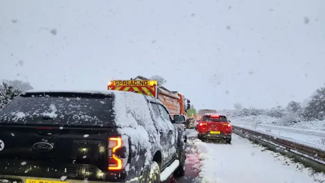 Photo shows several cars queuing on the A30 with heavy snow all around. An orange gritting lorry can be seen in front of a black truck.