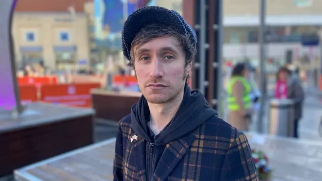 Joe Cook is wearing a hoodie and a chequered coat. He is wearing a flat cap. The background is blurred