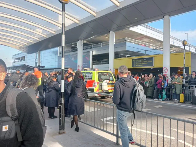 A big group of people stood on either side of the road at Gatwick Airport. A police vehicle drives through.