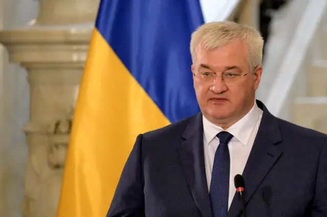 Ukraine's foreign minister Andriy Sybiga standing at a microphone, pictured from chest height up, wearing a dark suit, with a Ukrainian flag behind him and a marble column