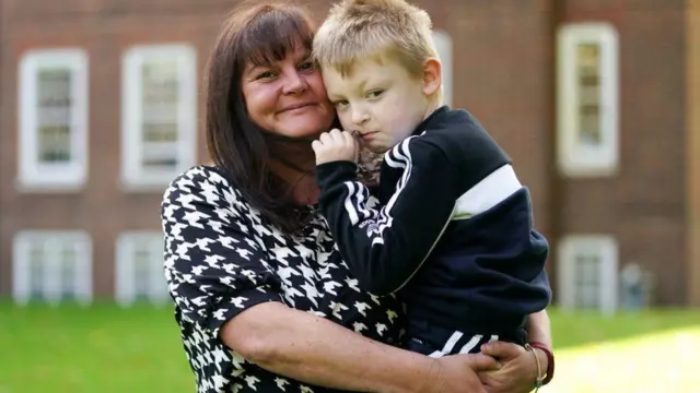 A woman with brown hair wearing a houndstooth top holds a young boy with blond hair in her arms - he is wearing a black tracksuit with white stripes. The background is blurred but red-brick buildings and grass are visible.