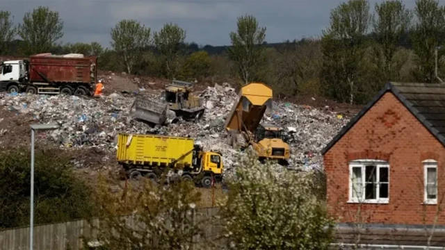 A view of a landfill site, with garbage lorries on the site. Next to the quarry is a red-brick house, a fence, and a few small trees.