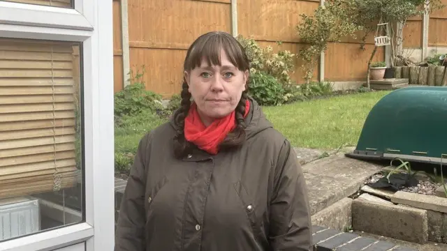 A woman with brown hair in two plaits wears a brown coat and red scarf. She looks at the camera with a straight face while standing in a residential garedn. An open door can be seen to the left of her.