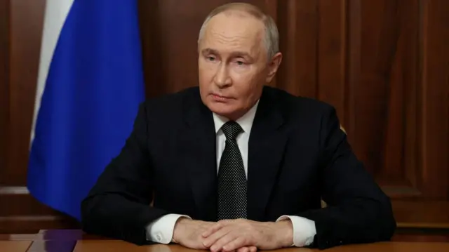 President Putin dressed in a dark suit and tie, sat with his hands folded in front of him on a desk during a television address