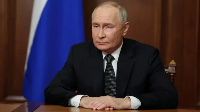 Russian president Vladimir Putin sat behind a desk. He has a Russian flag behind him.