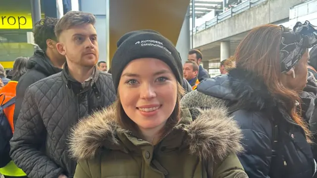 A woman in a green jacket and black hat in a queue.