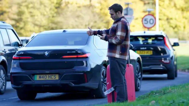 A man stands at the roadside with their thumb out in an attempt to hitchhike