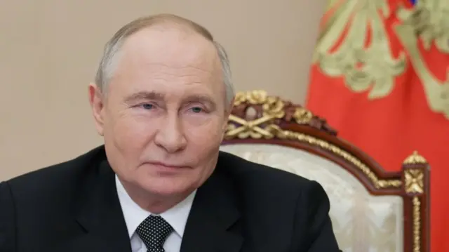 Vladimir Putin mid-shot cutting off below shoulders. He's wearing a black suit, white shirt and polka dot pattern tie as he sits on a wooden chair with gold inlays and cream fabric cushion back