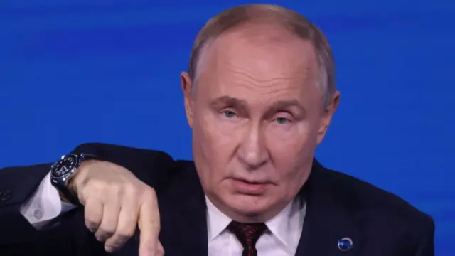 Mid-shot of Vladimir Putin cutting off just below the shoulders. He's in a black suit and white shirt, his right arm extended and his hand pointing down