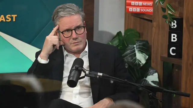 Keir Starmer sitting in front of a microphone with his fingers pressed against the right side of his temple, wearing an open-necked shirt and dark suit jacket