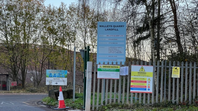 A light blue sign says 'Walleys Quary Landfill', attached to silver metal gates which has several other signs attached. To the left of the gates is a road and an orange and white traffic cone. Several trees are in the background.