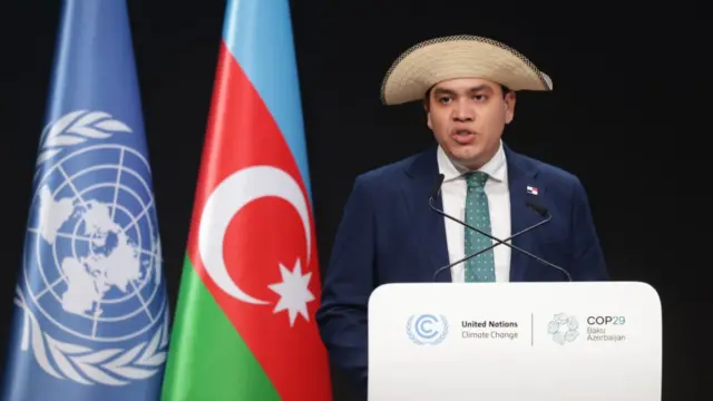 Juan Carlos Monterrey Gomez, wearing a large hat, making a speech in front of the Azerbaijan flag