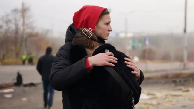 A woman in a black coat with a red hood hugs another woman, whose face cannot be seen.