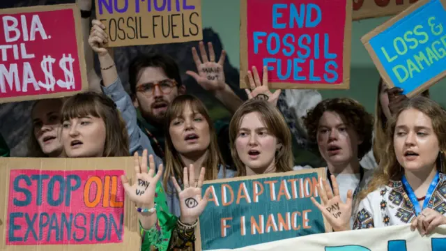 Climate activists protest at COP29 in Baku. In foreground, a young woman holds up her right hand showing the drawing of an eye on the palm of her hand. A left hand with the writing "pay up" on the palm is visible at the bottom left of the image