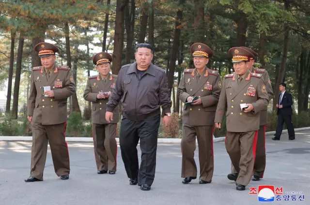 North Korean leader Kim Jong Un in middle of photo, surrounded by five men in military uniforms, walking down a path with tall trees in background