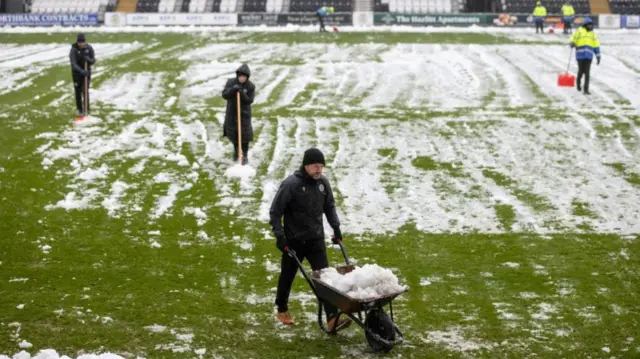 Staff clear snow from the pitch with shovels. One member of staff wheels away snow in a wheelbarrow.