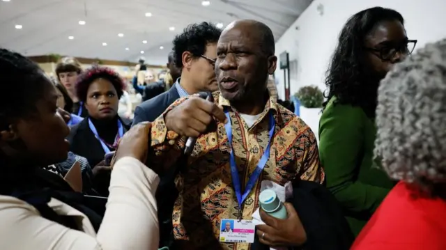 Evans Njewa, chair of the Least Developed Countries group, standing in a crowd, holding a microphone. A woman is holding his right forearm which is raised.