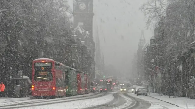 A row of buses lined up in the city centre. It is snowing heavily.