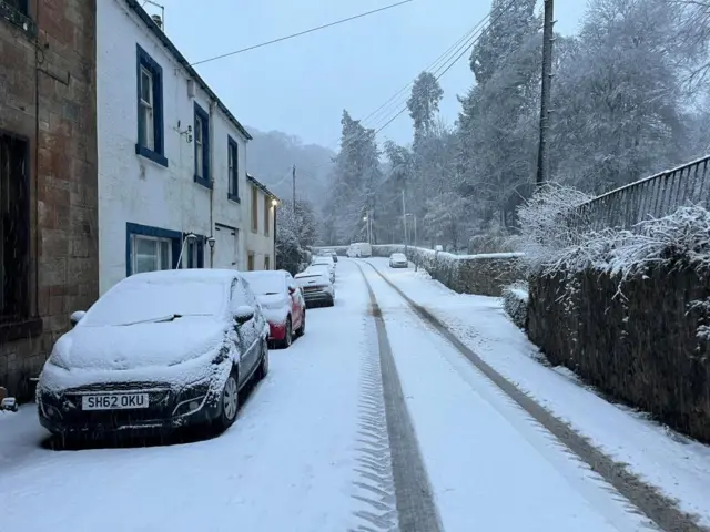 Snowy street scene. Row of houses on leftside, with street on right covered in snow with car tracks, parked cars covered in snow and tall trees in distance covered in snow