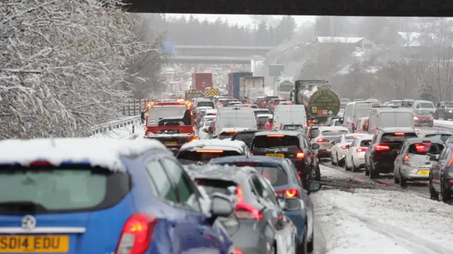 A view from a car of a hugs traffic jam in snowy conditions where a sliproad meets the M80 motorway. All the cars are showing their break lights.